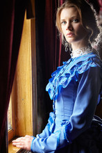 Young Woman In Blue Vintage Dress Standing Near Window In Coupe
