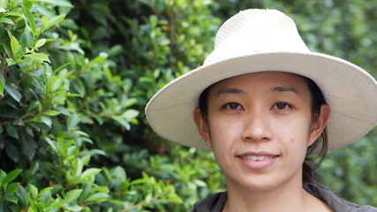 Wall Mural - Video of Beautiful smiling Asian woman closeup portrait. Wearing hat with natural green bush background. modern life condominium