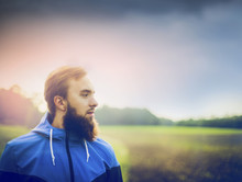 Young Man With A Beard Wearing A Blue Jacket And In Profile Against Green Field And Sky