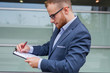 © jakubzak - Portrait of young businessman with beard standing in front of office building.