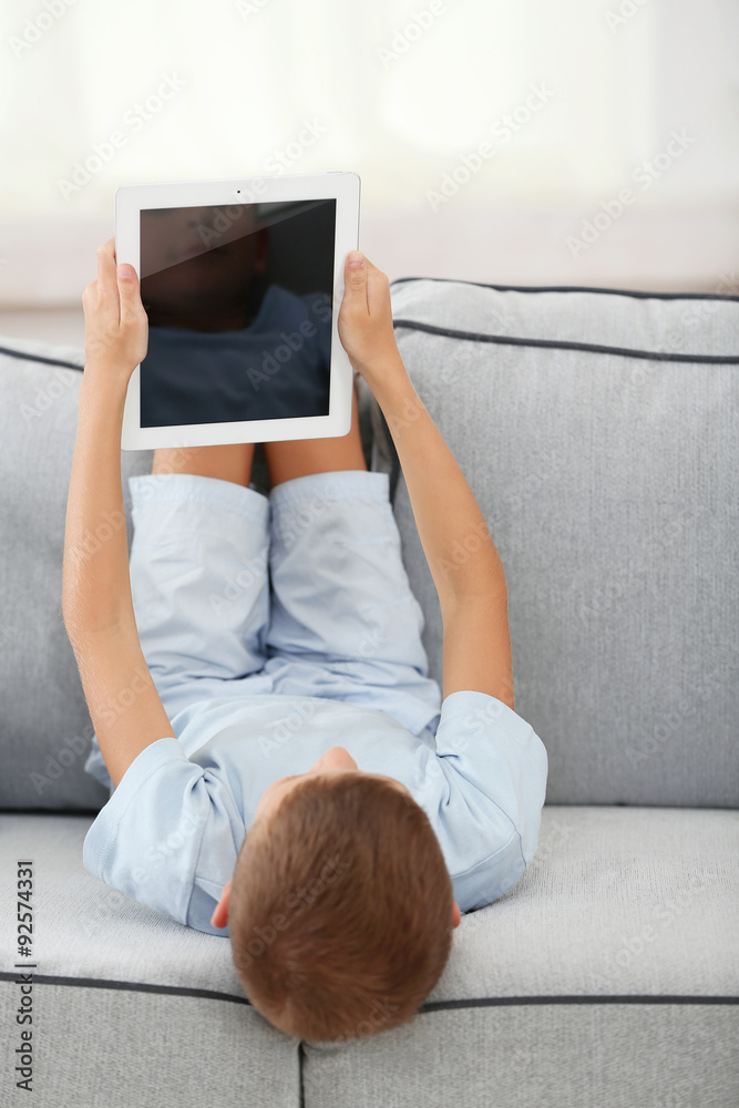 Little boy with digital tablet sitting on sofa, on home interior background