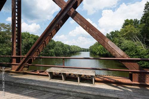 Fotografia, Obraz Bench on the Wallkill River Rail Trail Bridge in Upstate New York with a view of the river through the bridge structure