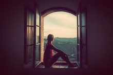 Young Woman Sitting In An Open Old Window Looking On The Landscape Of Tuscany, Italy.