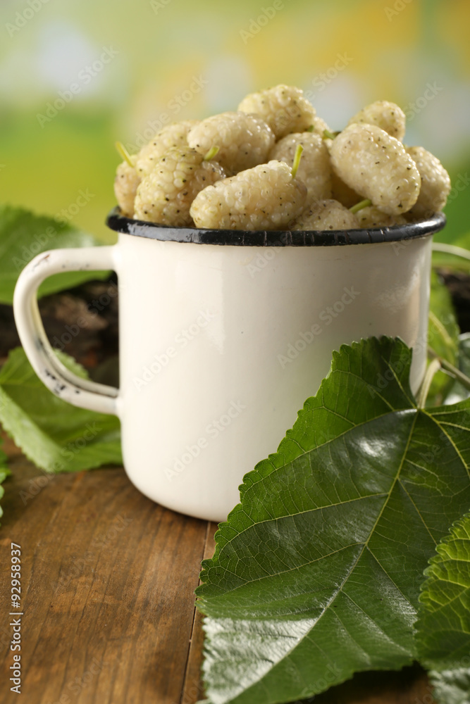 Ripe mulberries in mug with green leaves on table close up