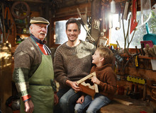 Three Generations - Father, Grandfather And Son Are Working On A Wooden Bird House. They Are In The Grandfather Workshop, He Is Wearing A Green Apron And Several Tools Are Hanging From The Ceiling