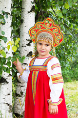 Wall Mural - little girl in Russian national a sundress and a kokoshnik stand near a birch in summer day