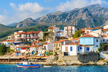 Typical Greek Fishing Boat In Kokkari Bay With Town Houses In Background, Samos Island, Greece