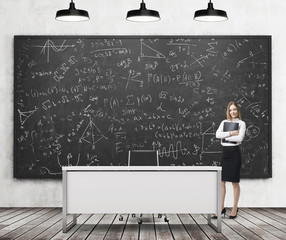 A beautiful girl presents math calculations on the black chalk board in a modern classroom. Three black ceiling lights, wooden floor and concrete wall. A concept of business education.