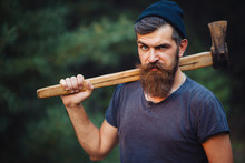 Brutal Brunette Bearded Man In Warm Hat With A Hatchet In The Woods On A Background Of Trees