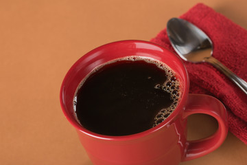 Freshly poured black coffee in a red mug on an amber brown backdrop with spoon and napkin on the side