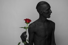 Gothic And Halloween Theme: A Man With Black Skin Holding A Red Rose, Black Death Isolated On A Gray Background In Studio