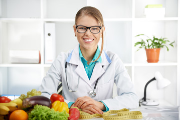 Smiling female dietitian sitting at the table with colorful fruits and vegetables in clinic. Concept of diet, lose weight and healthcare.  