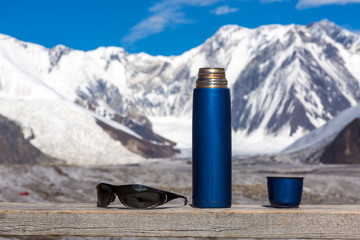 Blue Travel Thermos with Opened Cup and Sunglasses on Wooden Table High Mountains on Background