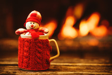 Cup Of Hot Drink In Front Of Warm Fireplace. Holiday Christmas Concept. Mug In Red Knitted Mitten, Decorated With Snowman Toy, Standing Near Fireside. Cozy Relaxed Magical Atmosphere In A Chalet.