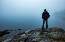 Man Standing On Cliff Looking Out Over Fog Covered Lake