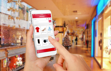 Wall Mural - Smart phone online shopping in woman hand during Christmas. Shopping center in background. Buy clothes shoes accessories with e commerce web site