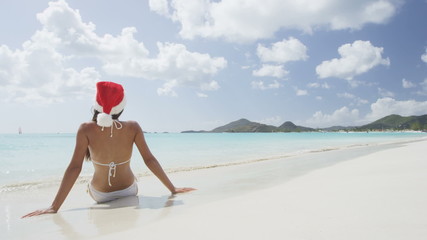 Wall Mural - Christmas beach vacation travel woman wearing Santa hat and bikini enjoying winter holidays getaway on tropical beach. Multicultural Asian Chinese / Caucasian model happy sitting in sand. RED EPIC.