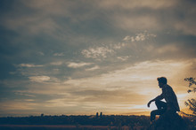 Silhouette Of Young Man Sitting On Sunset Or Sunrise. Confident Teenage Boy Thinkig On Cliff Stone. Hope. Sadness. Freedom.