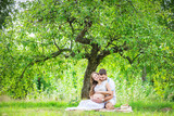Fototapeta  - Happy young couple expecting baby, pregnant woman with husband touching belly, sitting on green grass 