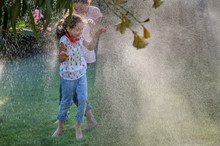 Two Sisters Having Fun Playing With Water From A Sprinkler In The Garden