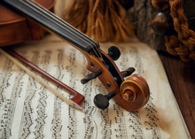 Old Wooden String Instrument (violin) Resting On Classical Sheet Music. Focus Is On The Scroll, Tuning Pegs, Bow, And Part Of The Neck.
