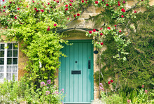 Green Wooden Doors In An Old Traditional English Stone Cottage Surrounded By Climbing Red Roses And Flowers