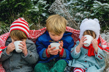 Three Children Wrapped In A Blanket Sitting In The Snow Drinking Hot Chocolate