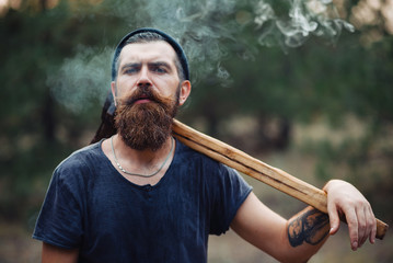 Lumberjack brutal bearded man in warm hat with a cigarette and an ax in the autumn forest at sunset