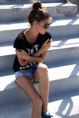 Wall Mural - Young European teenage brunette curly model posing on stadium concrete stairs, wearing blue jeans mini shorts, sneakers, round sunglasses and black t-shirt, ready to party