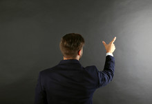 Back View Of Caucasian Young Man In Navy Blue Suit Pointing On Dark Grey Background