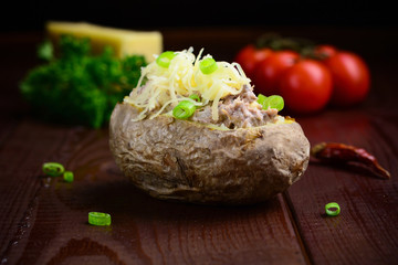 Baked potato stuffed with tuna and cheese garnished with green onion on dark brown wooden table. Low key photography