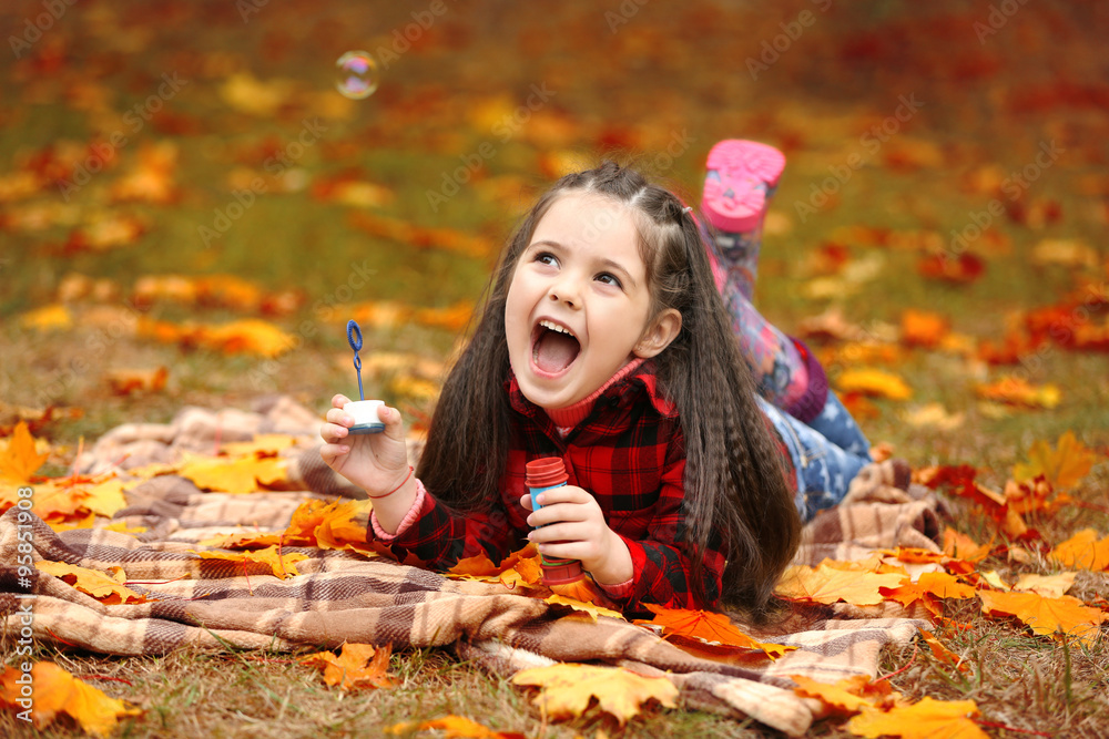 Cute girl lying on plaid and blowing soap bubbles in park
