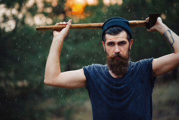 Wall Mural - Brutal brunette bearded man in warm hat with a hatchet in the woods on a background of trees