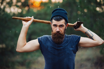 Canvas Print - Brutal brunette bearded man in warm hat with a hatchet in the woods on a background of trees