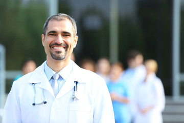 Canvas Print - Happy doctor with medical stuff behind standing against clinic entrance