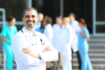 Canvas Print - Happy doctor with medical stuff behind standing against clinic entrance