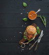 Fresh homemade burger on dark serving board with spicy tomato sauce, sea salt and herbs over black wooden background. Top view