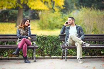 Wall Mural - Two young people sitting on benches in a park and talking on their mobile phones