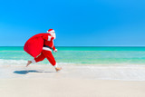 Fototapeta Miasto - Santa Claus running along beach with sack full of gifts