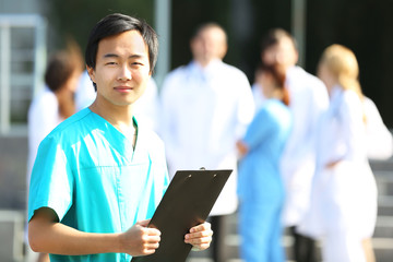 Poster - Young smiling doctor with clipboard in hands standing against group of medical workers