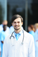 Canvas Print - Smiling doctor with medical stuff behind standing against clinic entrance