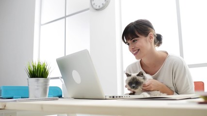 Sticker - Smiling woman at home typing on a laptop and social networking, she is holding and cuddling her cat