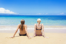 Two Women Sun Tanning On A Sunny Beautiful Beach. View From Behind Of Women Relaxing While On A Beautiful Island Vacation