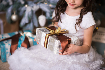 girl with curls and a smile sitting by a Christmas tree with a g