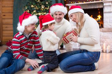 Wall Mural - Happy family in red hats with gift sitting at Christmas tree near fireplace