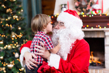 Wall Mural - Child toddler boy kissing Santa Claus sitting near Christmas tree