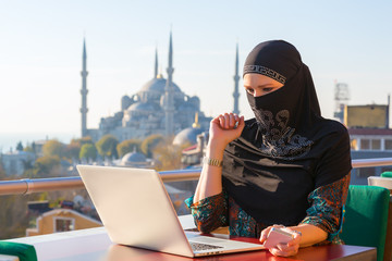 Wall Mural - Traditionally dressed Muslim Woman working on computer at outdoor balcony with oriental urban landscape on background