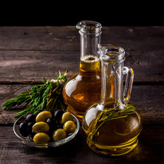 Two  bottles of olive oil, olive in a bowl and herbs on a wooden table.