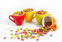 Plenty Assorted Color Dragees In Four Small Bright Colored Cups, The Orange Cup Is Felt Over The Sweets Are Spilled Over The White Table