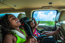 Brazilian Girls Singing And Laughing Sitting On Backseats In Car. Travelling Female Children In Child Safety Seat With Seatbelt On A Summer Day In Tropical Area